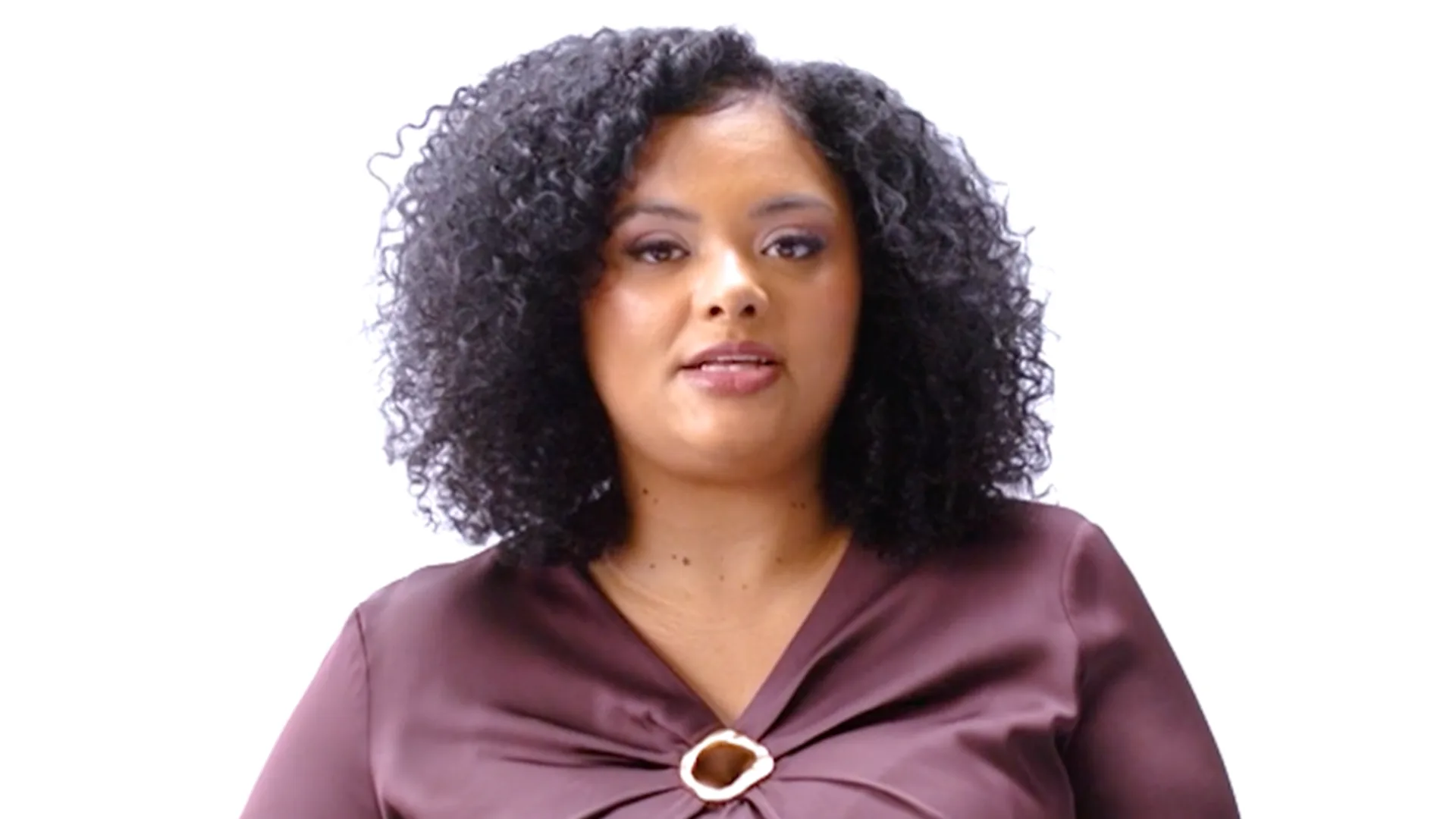 A Black woman with curly shoulder length hair wearing a deep plum blouse, looking directly at the camera against a white background.