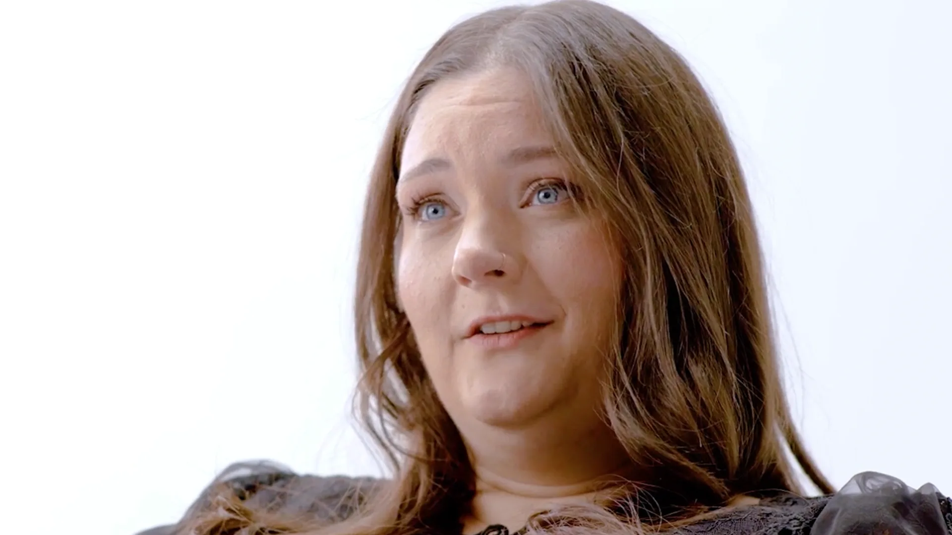 A portrait of a White woman with long brown hair speaking about how hidradenitis suppurativa (HS) impacts their daily lives , captured in a candid moment against a white background.