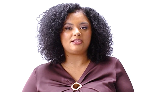 A Black woman with curly shoulder length hair wearing a deep plum blouse, looking directly at the camera against a white background.There is a green play button layered on top of the image 