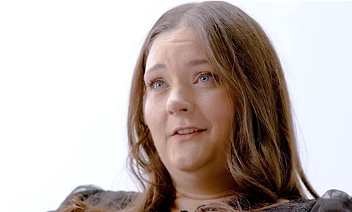 A portrait of a White woman with long brown hair speaking about how hidradenitis suppurativa (HS) impacts her daily life, captured in a candid moment against a white background. There is a green play button layered on top of the image 